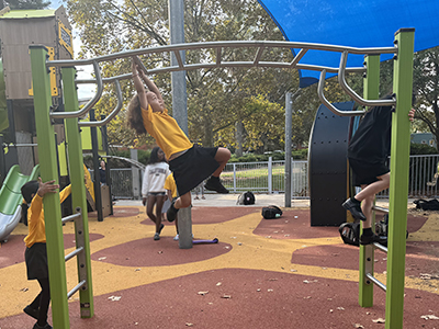 Three children wearing black and yellow school uniforms play on new green and silver monkey bars at the Alison Park Playground in Randwick. One girl watches from the ground, while another, with long, curly brown hair hangs from a bar with both hands looking toward the next bar. The third child is climbing down the other side.