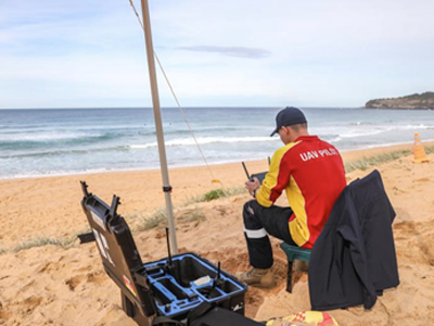 A UAV pilot sits on the beach, operating a drone under a shadecloth