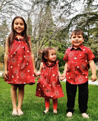 Three young children standing in the grass, wearing matching outfits featuring traditional Aboriginal art patterns