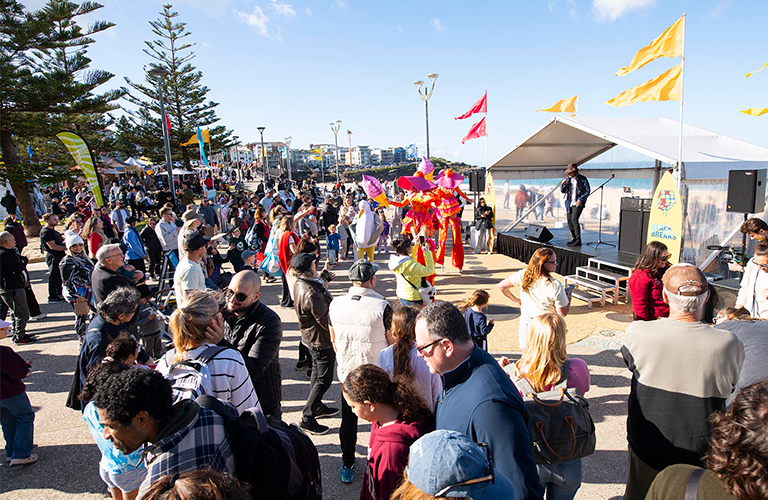 The Australian Surfing Walk of Fame presentation was held at Beach Breaks on Sunday 20 July 2025.. A photo of people gathering around a stage and roving performers on Maroubra Beach promenade.
