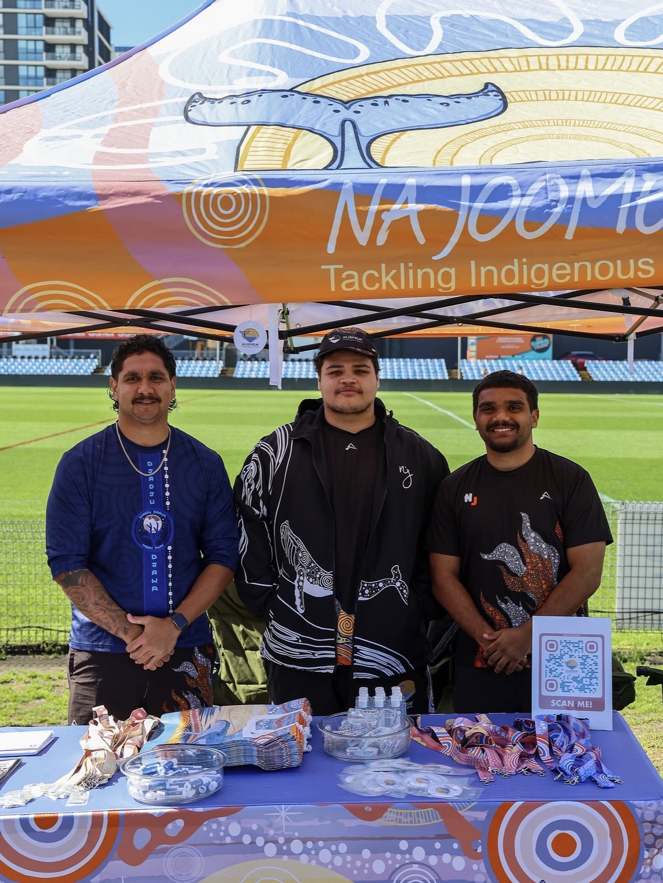 A group of young men standing under a branded marquee for the organisation "Na Joomelah"